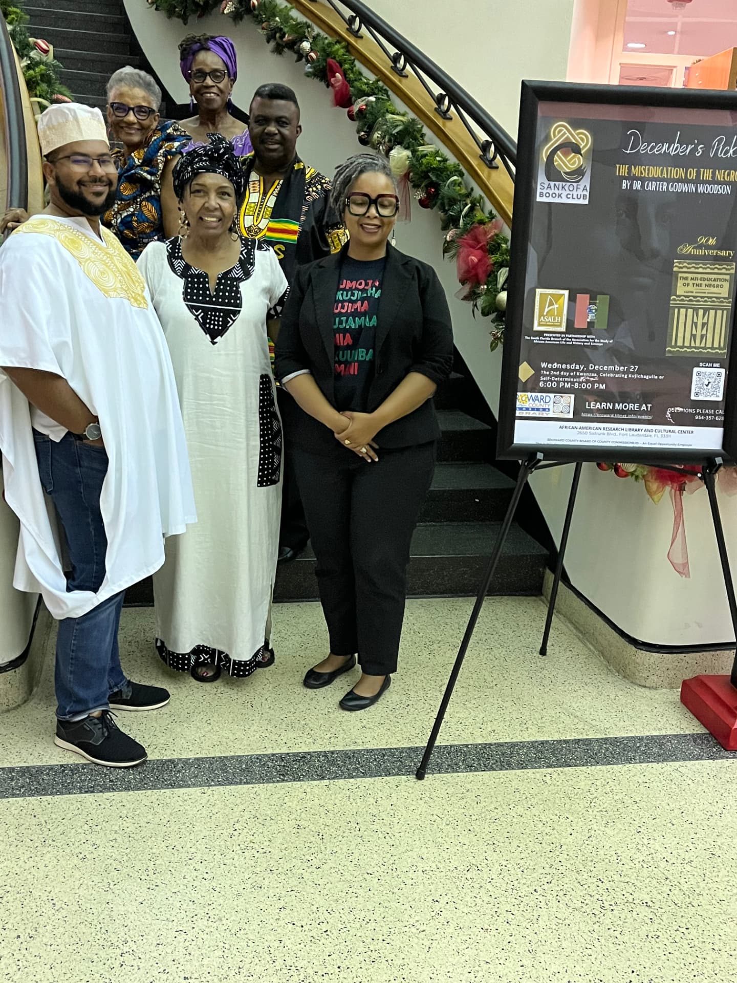 Six people standing near a grand staircase decorated with greenery and red bows, beside a Sankofa Book Club poster for a Kwanzaa event at the African-American Research Library and Cultural Center.