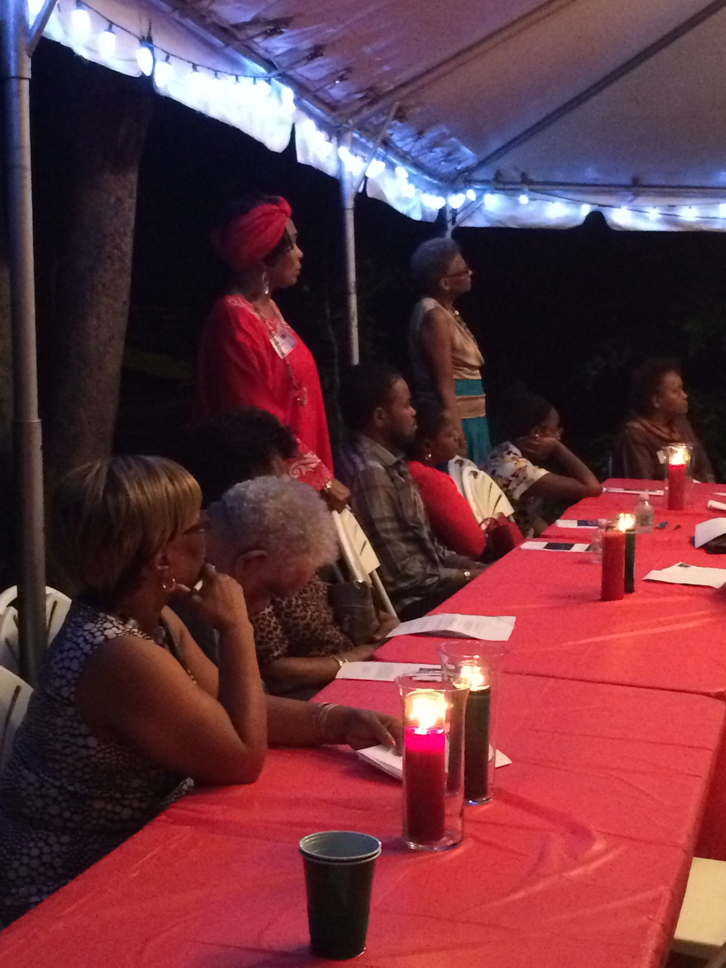 Guests seated at a long red-clothed table under a lit tent, with tall red and green Kwanzaa candles burning in glass holders.