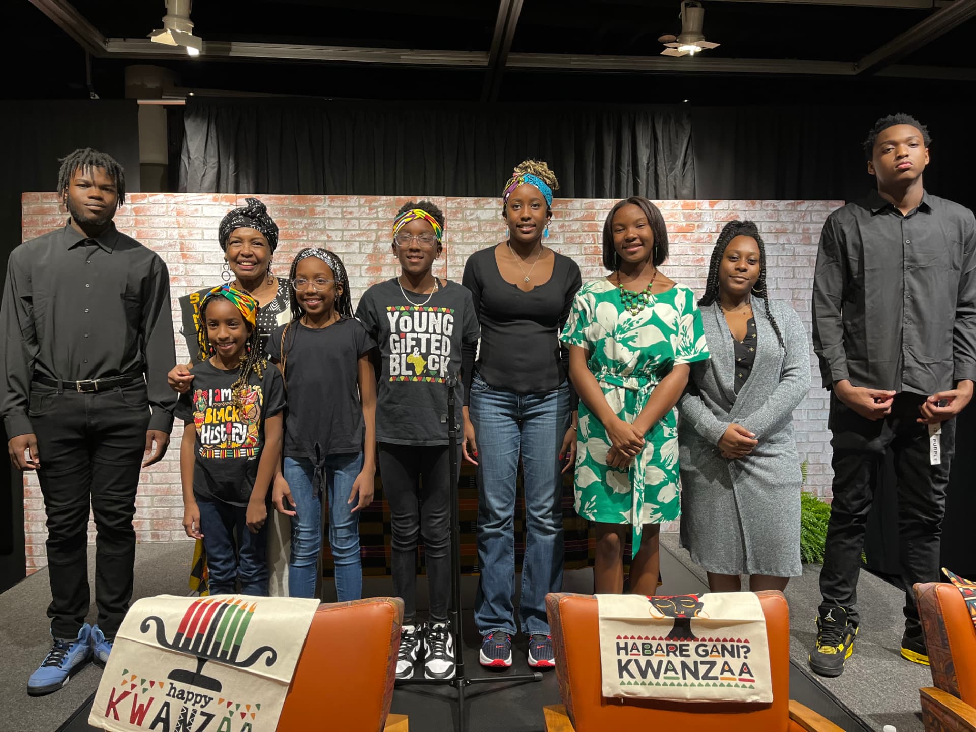 Nine people standing in a line on a stage with a faux brick backdrop, in front of two chairs draped with Kwanzaa covers reading Happy Kwanzaa and Habari Gani.
