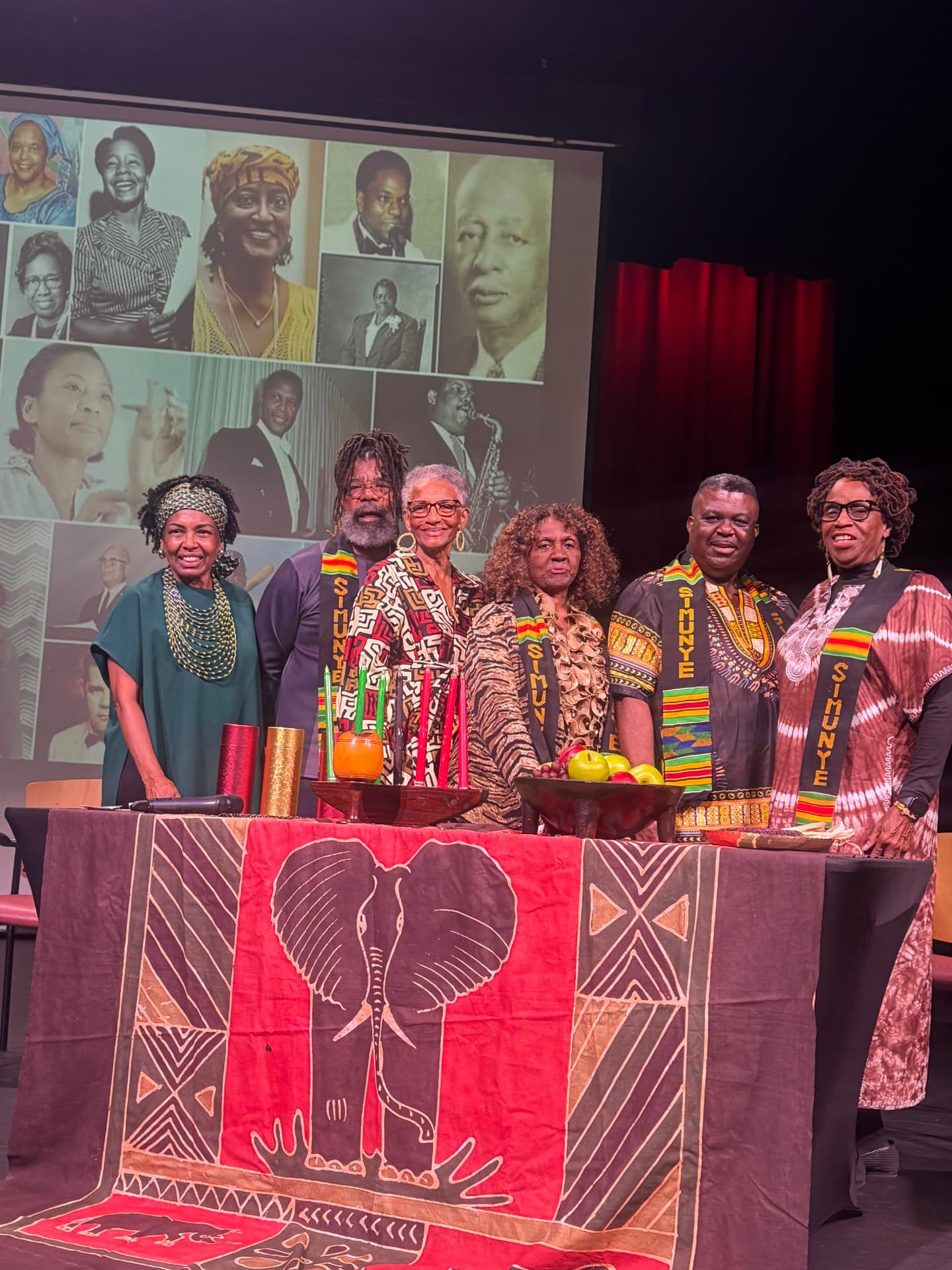 Simunye members wearing SIMUNYE sashes standing behind a decorated table with a kinara during a Kwanzaa celebration